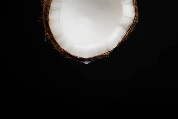 A drop of coconut water dripping from half a coconut on a black background, space for text.