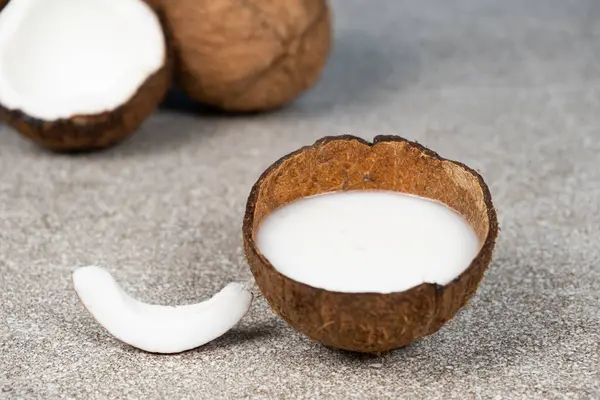Coconut milk in coconut shell and half coconut, chopped flesh on grey background.