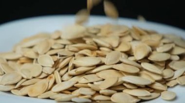Kabak çekirdekleri kabuksuz siyah arka planda yavaş çekimde bir tabağa düşüyor.