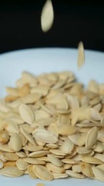 Kabak çekirdekleri kabuksuz siyah arka planda, dikey ağır çekimde bir tabağa düşüyor.