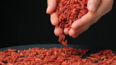 Ellerinden dökülen bir avuç kurutulmuş kırmızı kurt üzümü siyah arka planda ağır çekimde..