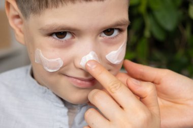 Annem bir çocuğun yüzüne yüz kremi sürüyor. Çocuklar için cilt bakımı ve korunma.