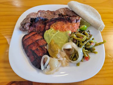 Lezzetli bir tabak ızgara sucuklu biftek, yanında guacamole, salsa, ızgara soğan ve nopal salata.