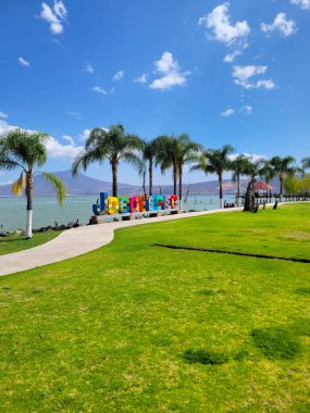 Canlı Jocotepec işareti, güneşli bir günde göl kıyısında yeşil çimenler ve palmiye ağaçları ile mavi gökyüzüne karşı duruyor.