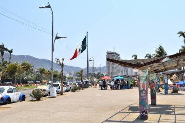 Acapulco, Meksika - 27 Nisan 2024: Arkasında bayrak dalgalanan işlek bir cadde. Etrafta dolaşan bir sürü insan ve cadde boyunca park etmiş arabalar var.