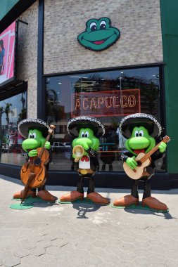 Acapulco, Mexico - April 27 2024: Vertical View of Three green statues of frogs musicians playing instruments in front of a store clipart