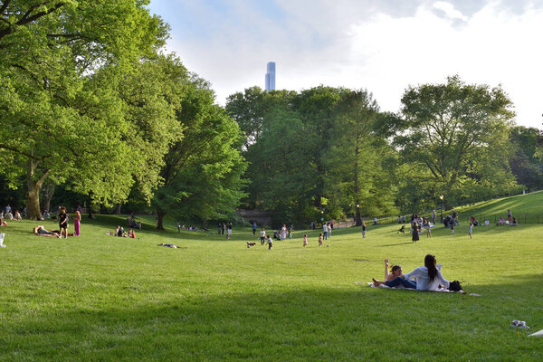 A large group of people are enjoying a sunny day in central park. There are many people sitting on the grass, some of them are reading books. NY
