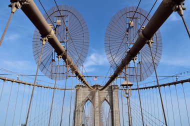 Brooklyn köprüsü, üzerinde iki büyük dairesel tekerleği olan büyük metal bir yapıdır. Gökyüzü açık ve mavi. Köprünün üzerinde dalgalanan bir bayrak var.