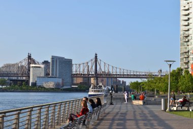 New York, ABD - 18 Mayıs 2023: Bir grup insan nehir kenarındaki banklarda oturuyor, New York şehri ve köprü manzarasının keyfini çıkarıyorlar.