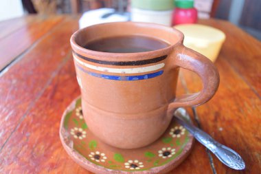 Bir fincan kahve yanında bir kaşıkla ahşap bir masada oturuyor. Bardakta çiçek deseni var ve üzerinde mavi bir şerit var. Kaşık gümüş ve fincanın kenarında duruyor.