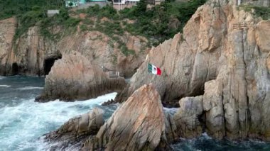Meksika bayrağının La Quebrada, Acapulco 'nun kayalıkları yakınındaki hava durağan görüntüsü. Aşağıda okyanus dalgaları çarpıyor.
