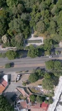 Şehir yapılarıyla çevrili ağaçlarla dolu bir şehir parkının güzelliğini yansıtan dikey bir dron tepsisi huzur veren yeşil bir alan sunuyor.