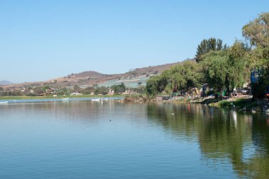 Meksika, Jalisco 'daki Cajititlan Gölü' nün sakin manzarası, yemyeşil bitki örtüsü ve açık mavi gökyüzünün altındaki yuvarlanan tepelerle çevrili.