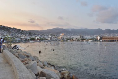 Acapulco, Meksika - 27 Nisan 2024: Günbatımındaki Acapulco Körfezi manzarası, şehir binalarını, dağları ve Otis Kasırgası 'ndan altı ay sonra suyun tadını çıkaran insanları gösteriyor