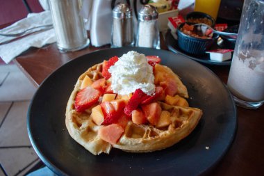 Günlük restoran ortamında siyah tabakta sunulan kremalı ve tropikal meyveli taze waffle kahvaltısı.