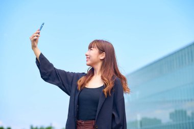 Mavi gökyüzünün altında akıllı telefon tutan genç bir kadın.