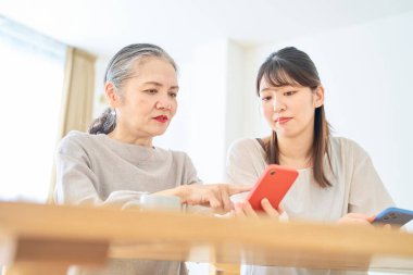 Son sınıf öğrencisi bir kadın ve genç bir kadın evde akıllı telefon ekranlarına endişeyle bakıyorlar.