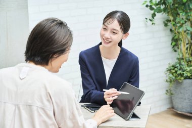 Takım elbiseli bir kadın, odada sade giyinmiş bir kadına açıklama yapıyor.