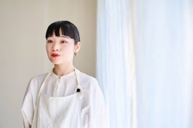 Önlüklü genç bir kadın odanın penceresinde mola veriyor.