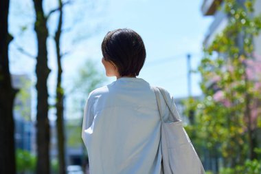 Güneşli bir günde alışveriş torbalarıyla şehirde yürüyen bir kadının arka görüntüsü.