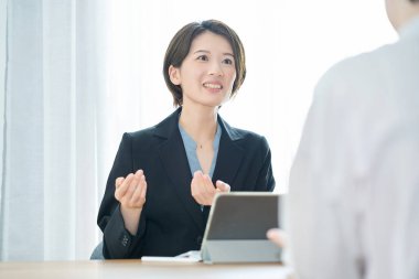 Takım elbiseli bir kadın gülümsüyor ve düz elbiseli bir kadına açıklama yapıyor.