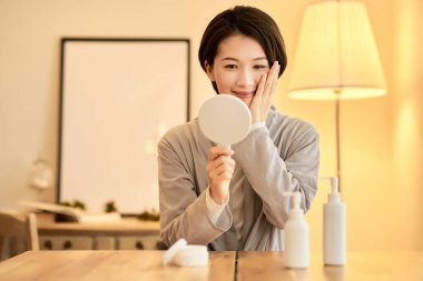 Sıcak ışıklandırmalı bir odada el aynasıyla kendini kontrol eden bir kadın.