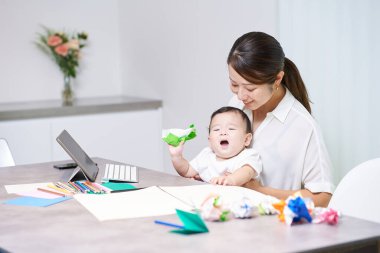 Bir kanepede uzaktan çalışan annesinin yanında oynayan bir çocuk.