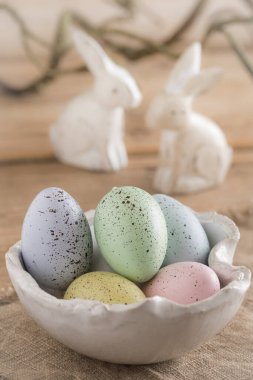 Dekoratif Paskalya yumurtaları pastel renklerde, beyaz el yapımı bir kâse içinde ahşap bir masada, arka planda iki beyaz tahta Paskalya tavşanı.