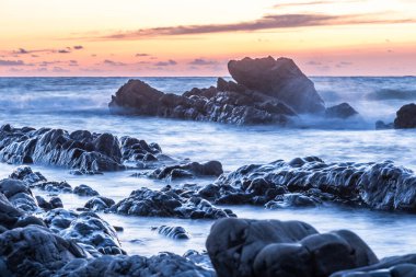 Gün batımında bir sahilin kayalık kıyısı, uzun süre maruz kalınca bulanık su
