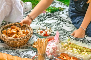 Çimenlerde çeşitli parmak yiyecekleriyle piknik yapan bir kadın eli, içinde ekmek olan bir sepet getirir.