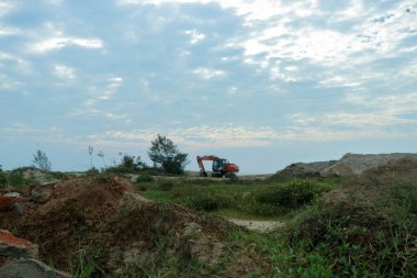 Altın bir gökyüzünün altında, ön planda yemyeşil yemyeşil bir kıyı manzarası yakınında çalışan parlak turuncu bir kazıcı..