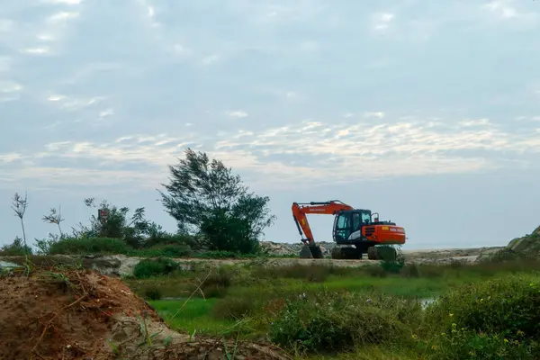 Altın bir gökyüzünün altında, ön planda yemyeşil yemyeşil bir kıyı manzarası yakınında çalışan parlak turuncu bir kazıcı..
