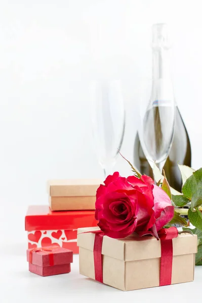 On a light background, a red rose and boxes with gifts, in the background two glasses and a bottle of champagne. Romantic vacation concept.