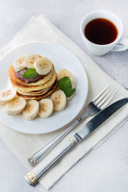 Işık masasında bir tabak krep, muz dilimleri, çatal bıçak takımı ve bir fincan çay var..