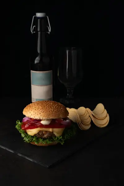 There is a burger on a dark background, a blurred image of chips and a bottle of beer in the background.
