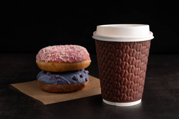 On a dark background there is a paper cup with coffee and two multi-colored donuts. Fast food concept.