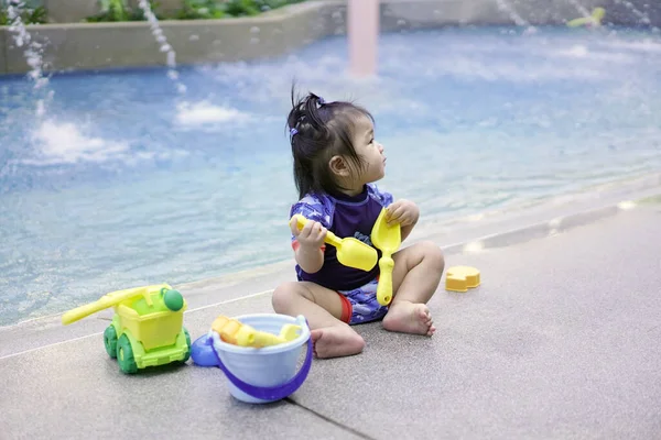Asian baby girl playing with water toys by the pool. Little girl playing in outdoor swimming pool on summer vacation on tropical beach island. Child learning to swim in outdoor pool of luxury resort. Water toy for kids