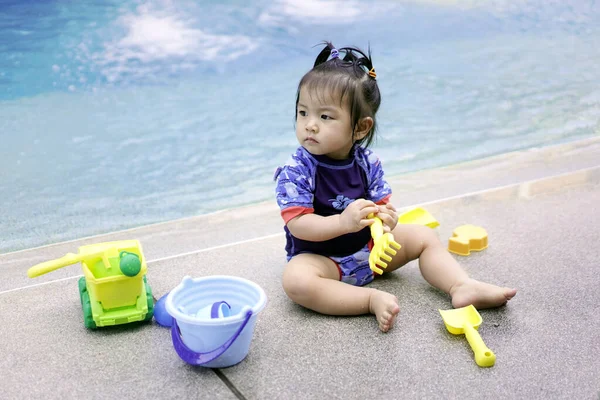 Asian baby girl playing with water toys by the pool. Little girl playing in outdoor swimming pool on summer vacation on tropical beach island. Child learning to swim in outdoor pool of luxury resort. Water toy for kids