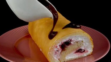 Pouring with chocolate sauce biscuit swiss roll with butter cream on black background rotate. Preparation sweet dessert on domestic kitchen. Slow motion. Close up.