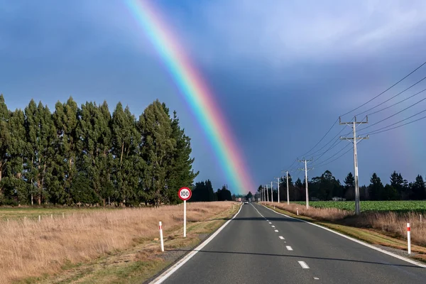 Photograph of a large rainbow over a regional road and agricultural field on the South Island of New Zealand