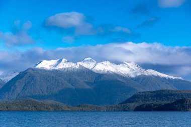Fiordland 'daki Te Anau' dan Yeni Zelanda 'nın güney adasındaki Manapouri' ye giderken büyük mavi bir göl ve karla kaplı bir dağ sırasının fotoğrafı.