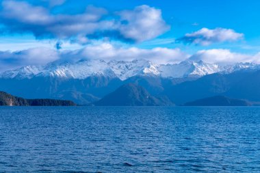 Fiordland 'daki Te Anau' dan Yeni Zelanda 'nın güney adasındaki Manapouri' ye giderken büyük mavi bir göl ve karla kaplı bir dağ sırasının fotoğrafı.