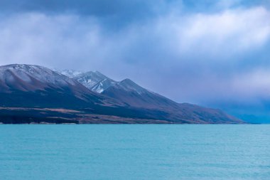 Pukaki Gölü 'nün fotoğrafı, bulutlu bir günde, arka planda kar örtülü dağlarla Yeni Zelanda' nın Güney Adası 'nda.