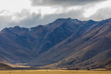 Yeni Zelanda 'nın Güney Adası' ndaki düşük seviyeli gri bulutların oluşturduğu büyük bir kahverengi tarım tarlasının arkasında uzanan kuru bir dağ sırasının fotoğrafı.