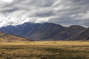 Yeni Zelanda 'nın Güney Adası' ndaki düşük seviyeli gri bulutların oluşturduğu büyük bir kahverengi tarım tarlasının arkasında uzanan kuru bir dağ sırasının fotoğrafı.