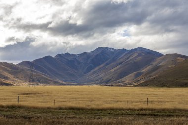 Yeni Zelanda 'nın Güney Adası' ndaki düşük seviyeli gri bulutların oluşturduğu büyük bir kahverengi tarım tarlasının arkasında uzanan kuru bir dağ sırasının fotoğrafı.