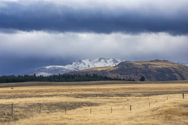 ニュージーランド南島の低レベルの灰色の雲を持つ大きな茶色の農場の背後を走る雪に覆われた山の範囲の写真 — ストック写真