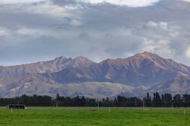 Yeni Zelanda 'nın güney adasında alçak seviyeli gri bulutların bulunduğu büyük yeşil bir tarla boyunca uzanan bir sıradağların fotoğrafı.