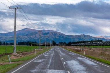 Yeni Zelanda 'nın güneyindeki düşük seviyeli gri bulutların bulunduğu kırsal bir alanda bir dağ sırasına doğru giden bir yolun fotoğrafı.