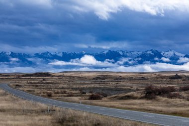 Yeni Zelanda 'nın güneyindeki düşük seviyeli gri bulutların bulunduğu kırsal bir alanda bir dağ sırasına doğru giden bir yolun fotoğrafı.